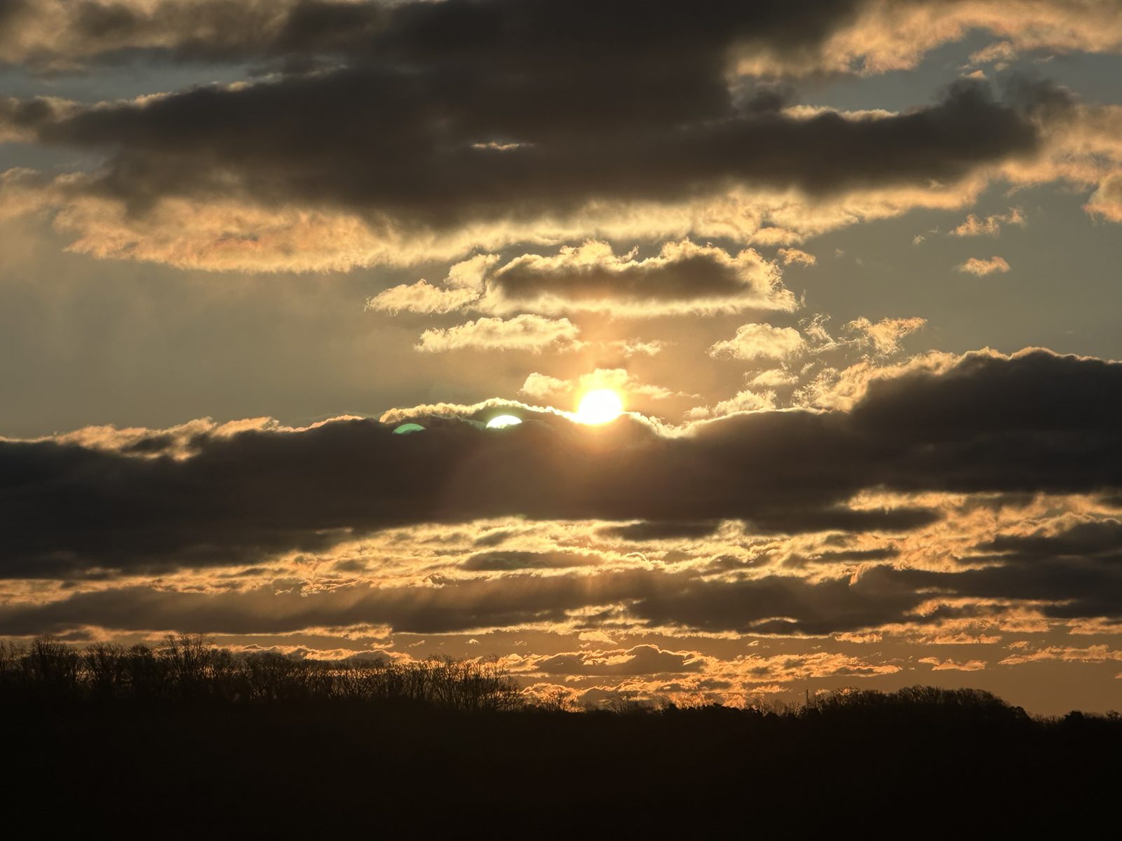 Sunset over a Vermont treeline, sun breaking through clouds with a green flash